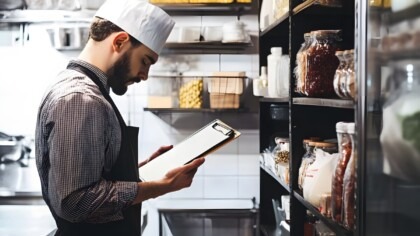 Owner checking inventory in restaurant kitchen, clipboard in hand, shelves of ingredients behind