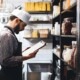 Owner checking inventory in restaurant kitchen, clipboard in hand, shelves of ingredients behind